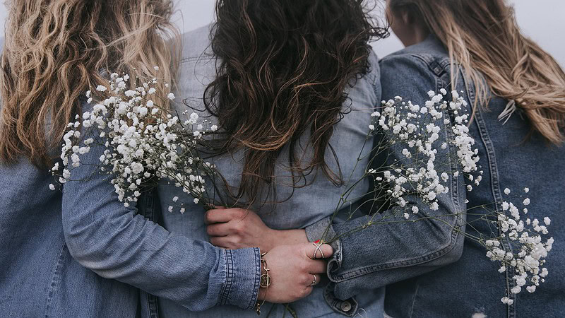 2 Frauen halten jeweils eine Blume in der Hand und haben diese auf dem Rücken einer 3. Frau liegen