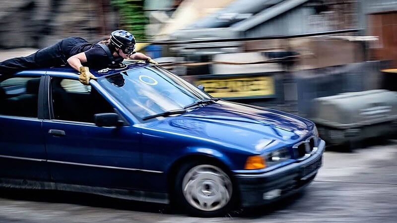 Ein behelmter Stuntman probt eine Szene auf dem Dach eines Autos