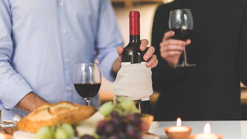 Ein Mann hält eine Rotwein-Flasche und ein Glas, eine Frau daneben trinkt den Rotwein