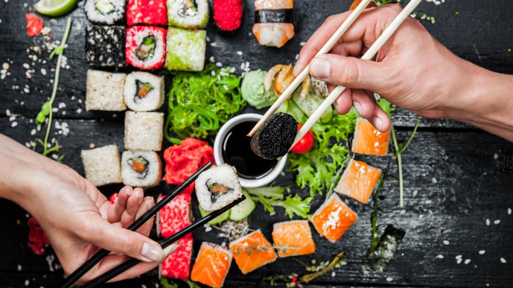 Großer dunkler Teller mit Sushi-Stücken und Toppings wie Ingwer und Wasabi, 2 Hände über dem Teller, die das Sushi mit Stäbchen halten