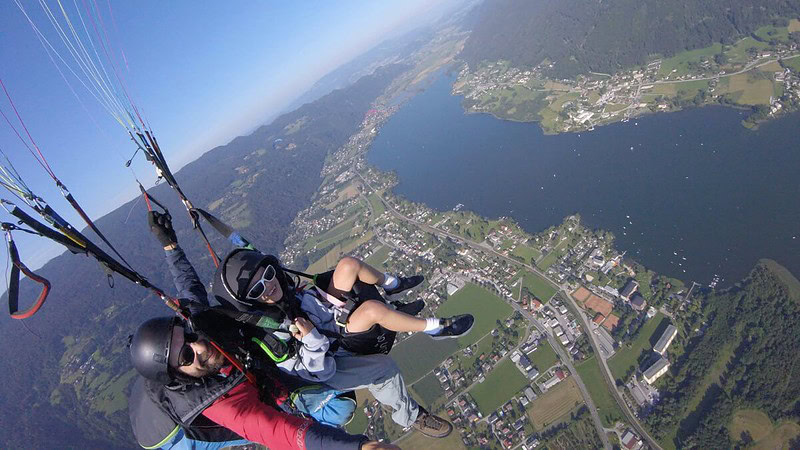 2 Personen fliegen zusammen mit einem Gleitschirm und gucken in die Kamera