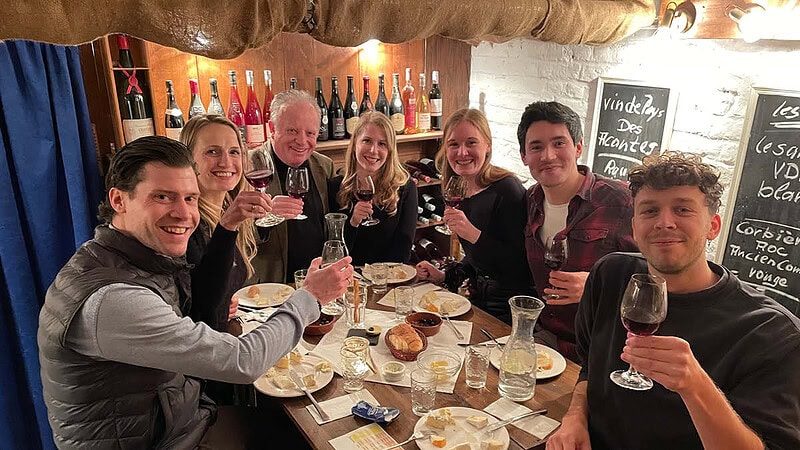 Eine Gruppe aus 7 Personen die mit einem Wein anstoßen, an einem gedeckten Tisch mit Käse Platten