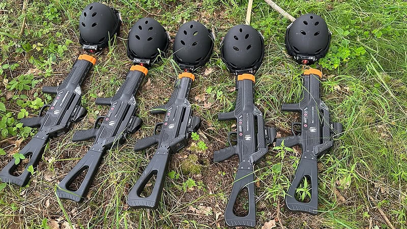 Fünf Lasertag Gewehre die in einer reihe nebeneinander liegen, mit fünf Helmen die über den Gewehren in einer Reihe liegen auf einer Wiese als Untergrund.