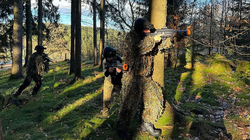 Lasertag Spieler mit Tarnkleidung die sich hinter einem Baum verstecken und in den Wald zielen.