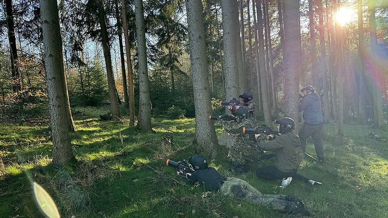 Fünf Lasertag Spieler mit Gewehren in der Hand die in Tarnkleidung gekleidet sind und alle in eine Richtung zielen. Sie befinden sich alle im Wald.