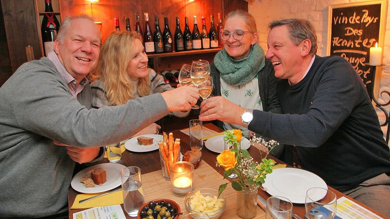 2 Paare die an einem Tisch mit Weinbeilagen sitzen und anstoßen.