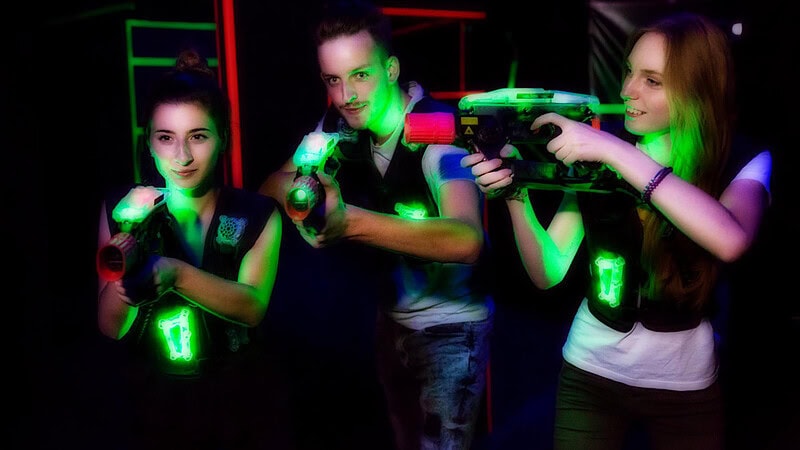 Zwei junge Frauen und ein Junger Mann die grün leuchtende Westen an haben und jeweils ein Lasertag Gewehr in der Hand halten.