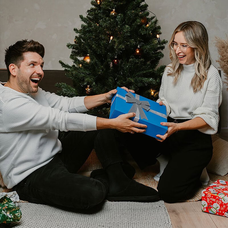 Paar übergibt die Geschenkbox der Erlebnisfabrik im Hintergrund ein Weihnachtsbaum